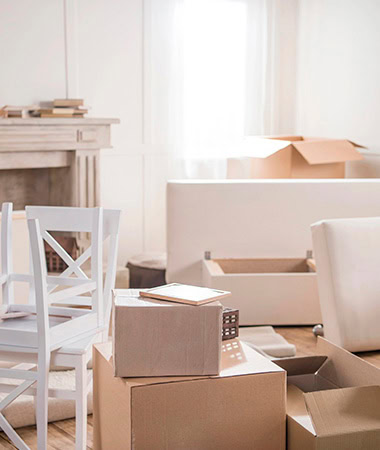 Una habitación con una silla blanca, cajas de cartón marcadas con 'Inicio' y varios artículos desempacados cerca de una ventana con cortinas transparentes.