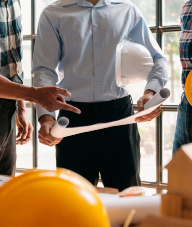 Un grupo de personas discutiendo planos arquitectónicos marca el inicio de un proyecto apasionante. Una persona sostiene un plano enrollado y un casco blanco mientras otros señalan los planos, indicando colaboración. Los cascos amarillos están colocados sobre una mesa en primer plano, listos para la acción.