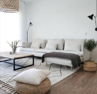 Nuevas promociones inmobiliarias en Palma de Mallorca con una sala de estar moderna y minimalista diseñada por Promotora cuenta con un sofá blanco, lámparas de pie negras, una mesa de centro de madera, un puf tejido y una manta gris sobre una alfombra blanca. Las paredes y los muebles son de tonos neutros.