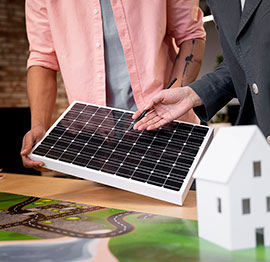 Dos personas sosteniendo un pequeño panel solar sobre una mesa con una casa modelo y un diseño de ciudad.