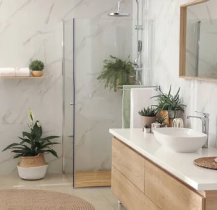 Un baño moderno con ducha con mampara de vidrio, tocador de madera con lavabo blanco, espejo de pared, plantas en macetas y un estante flotante con toallas. Perfecto para quienes buscan reformas integrales en Mallorca.