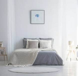 Un dormitorio minimalista con una cama cuidadosamente hecha con ropa de cama gris y blanca, una manta de crochet, dos almohadas, un cuadro enmarcado en la pared, una pequeña mesita de noche y una lámpara de pie insinúa reformas recién terminadas.